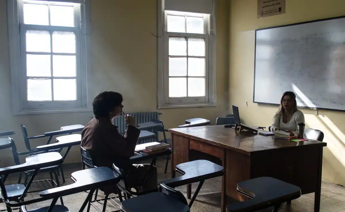 Aula con estudiantes en pupitres y docente explicando en la pizarra, luz natural por las ventanas.